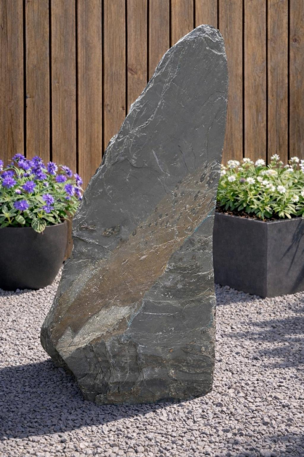 Grey slate monolith standing stone by 122 with natural textures, surrounded by pebbles and plants.