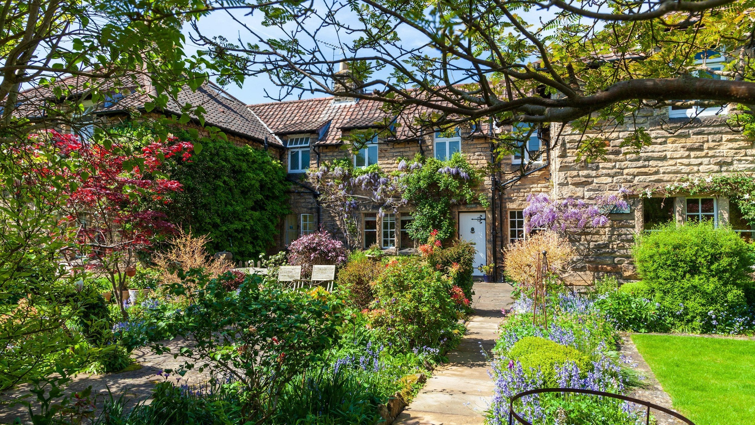 A stone pathway leads to a quaint stone cottage with wisteria vines, framed by vibrant flowers and lush greenery.