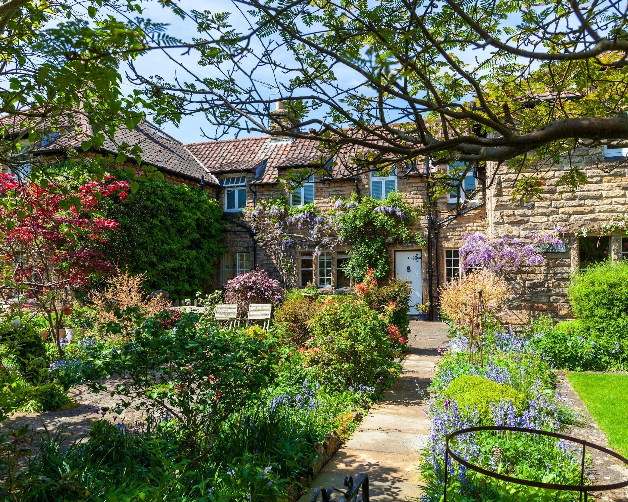 A quaint stone cottage with a lush garden in full bloom, showcasing colorful flowers and greenery along a stone path.