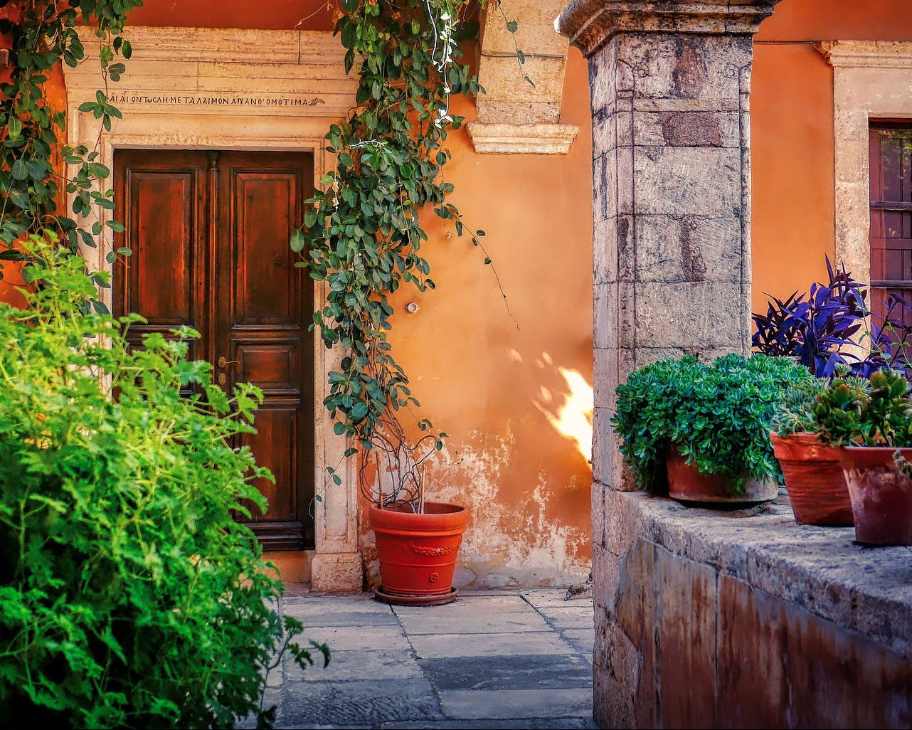 A sunlit courtyard scene with a wooden door, terracotta pots with plants, a stone pillar, and orange stucco walls. Greek text above the door.