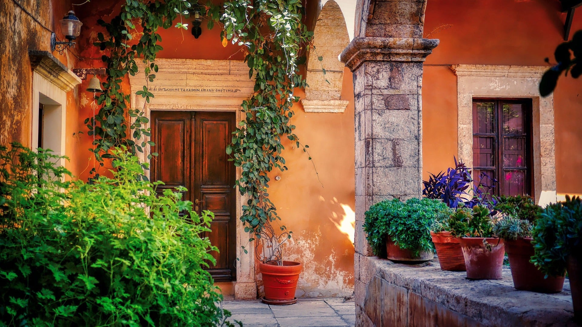 A sunlit Mediterranean courtyard with terracotta walls, a dark wood door, archway, and potted plants. Text above the door is in Greek. 
