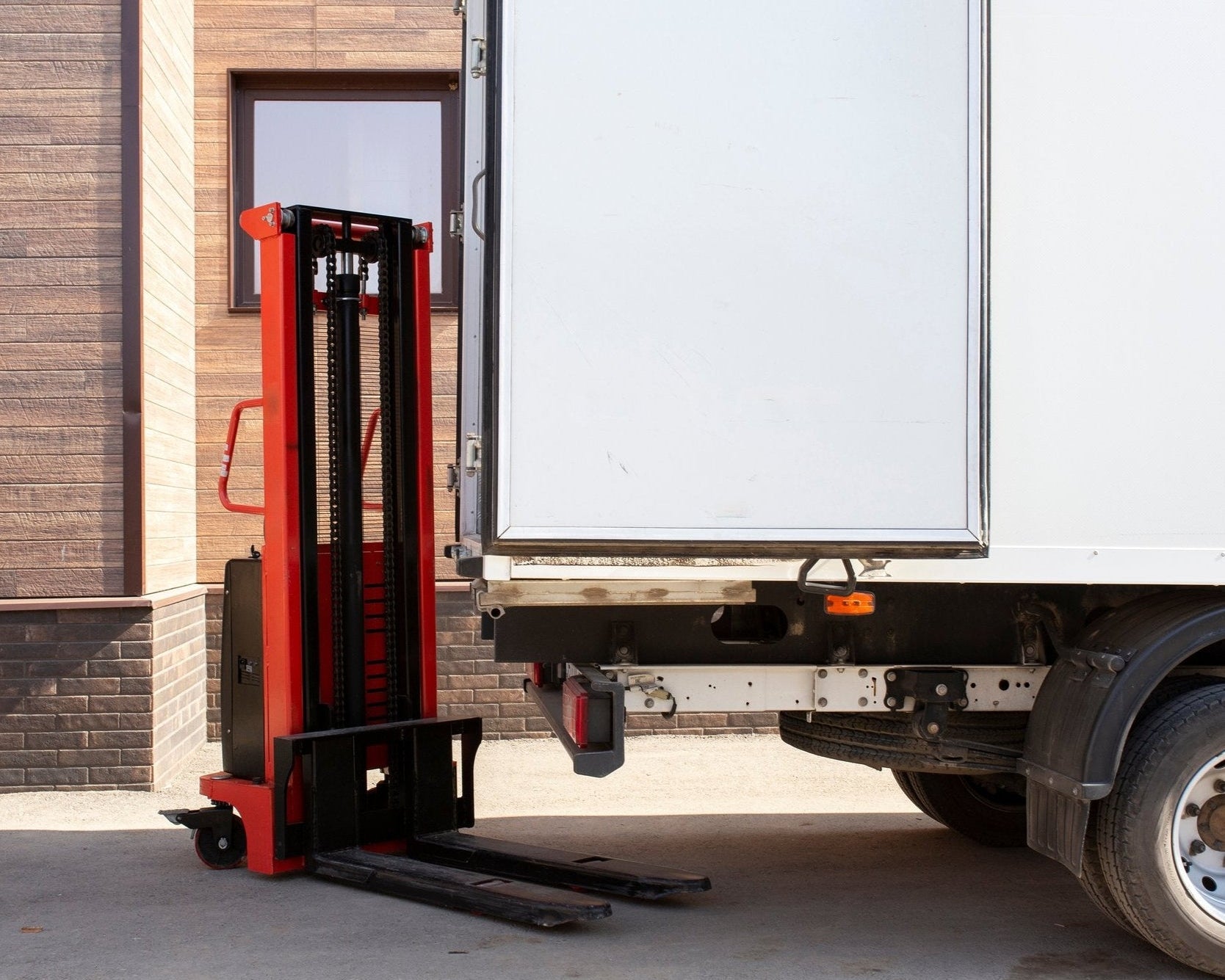 A red and black forklift sits next to the open back of a white truck in front of a building.