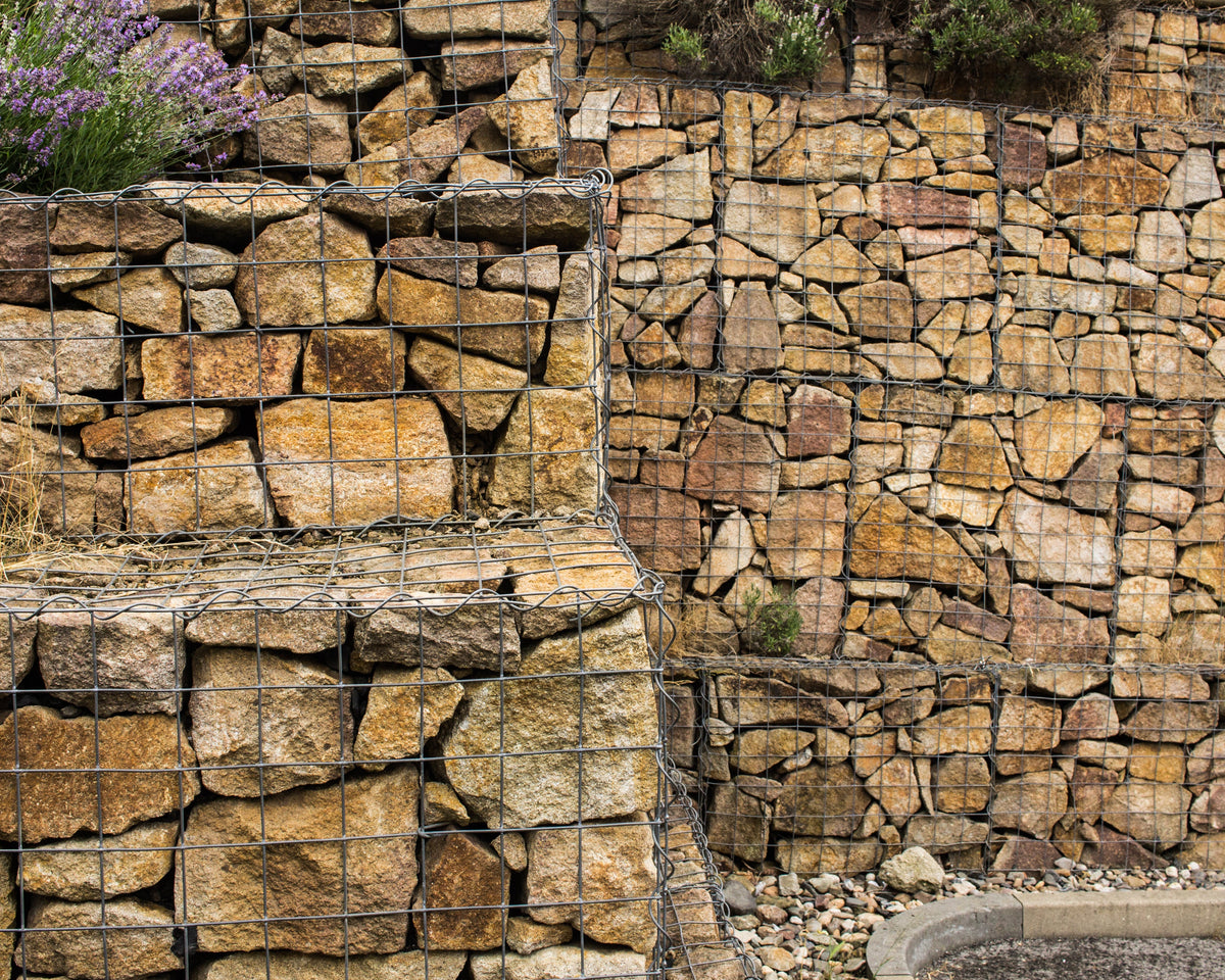 Gabion walls of stacked brown rocks enclosed in wire cages, with lavender growing atop one section. Neutral, outdoor setting.