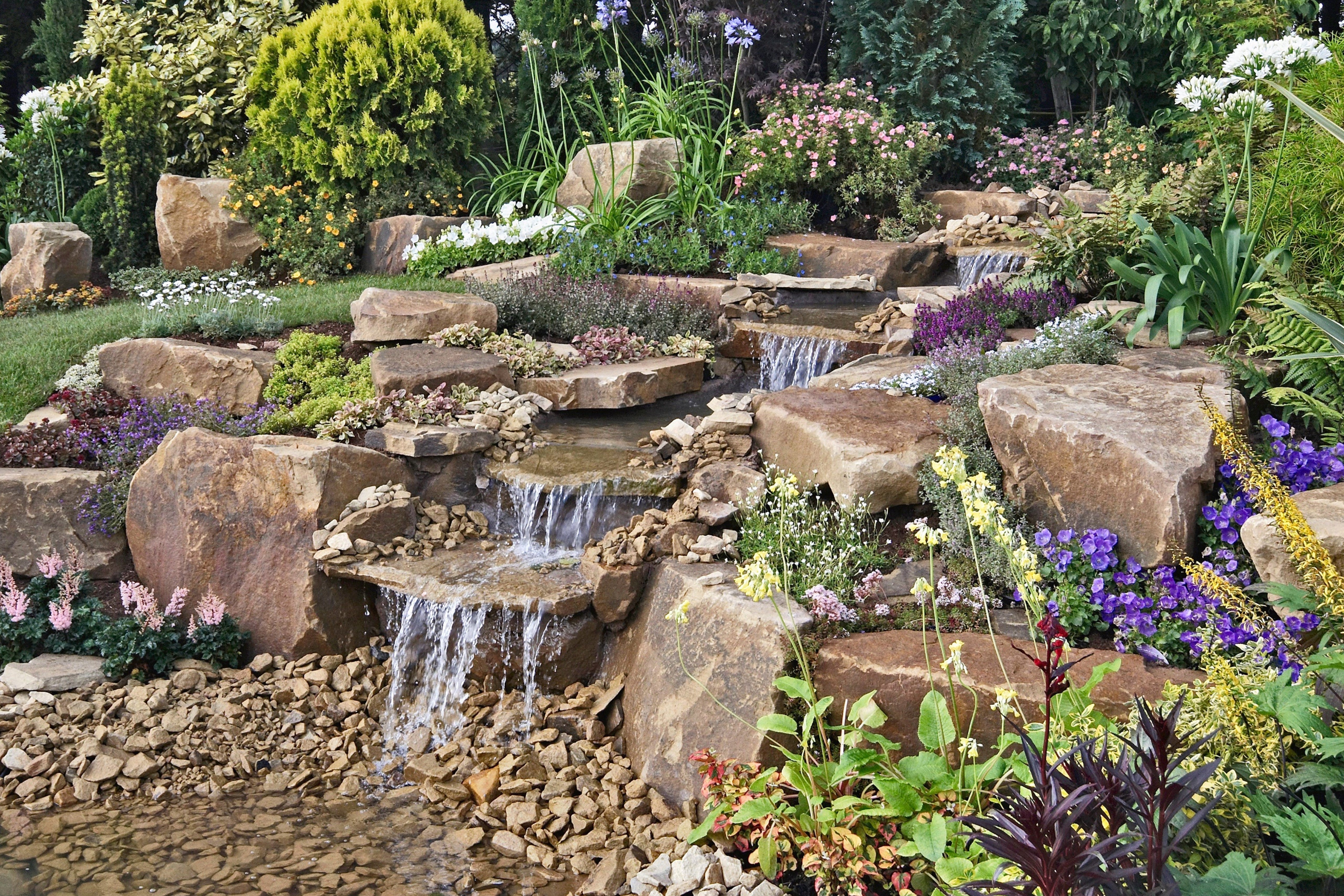 A multi-tiered rock garden waterfall surrounded by colorful plants and greenery creates a peaceful, natural atmosphere.