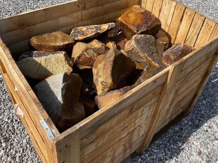 Golden granite rockery stones for gardens with dark, weathered accents in a wooden crate.
