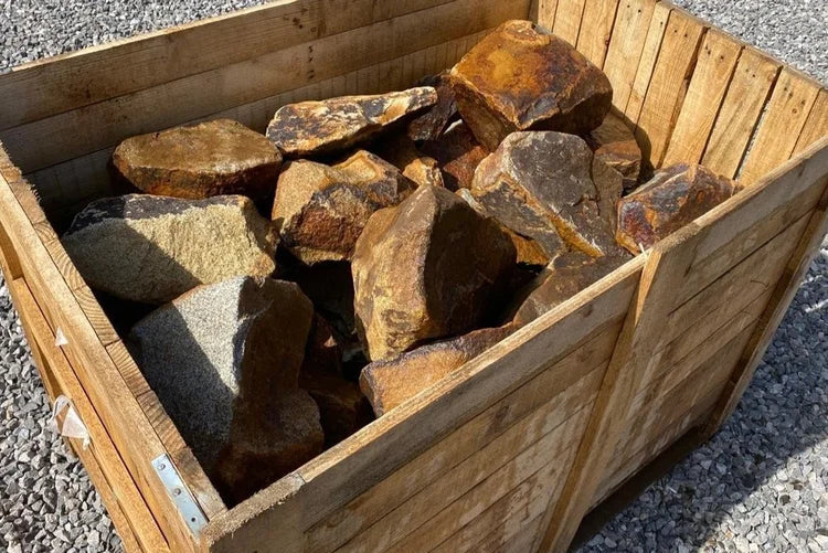 A wooden crate is filled with a pile of rough, brown and gray rocks of varying sizes. The crate sits on gravel.