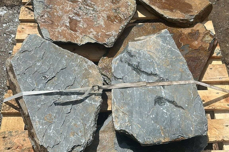 Rough-cut flagstone tiles in gray and brown are stacked on a wooden pallet and secured with a white plastic strap.