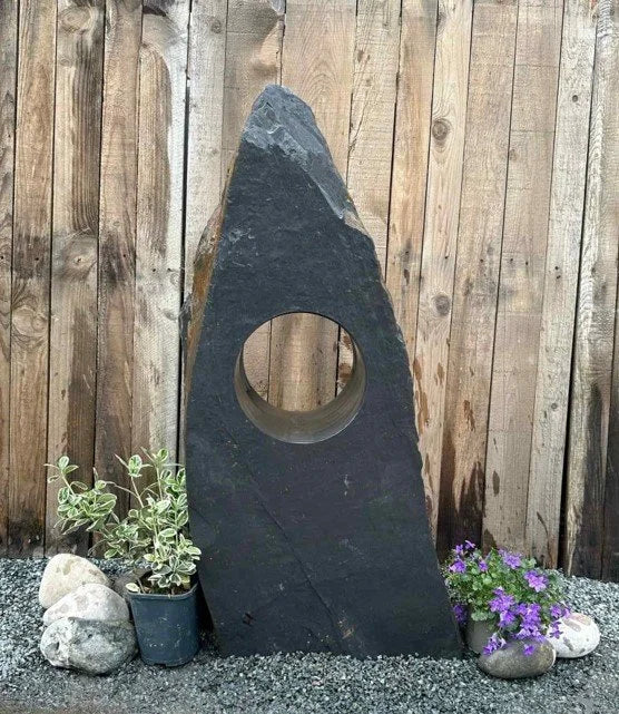 Grey slate Hollolith Water Feature by Hollolith, with a centered hole, sitting amongst gravel and plants.