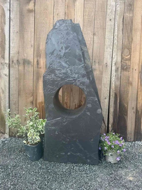 Hollolith grey slate water feature by Holloliths with a circular cut-out and plants on a gravel bed.
