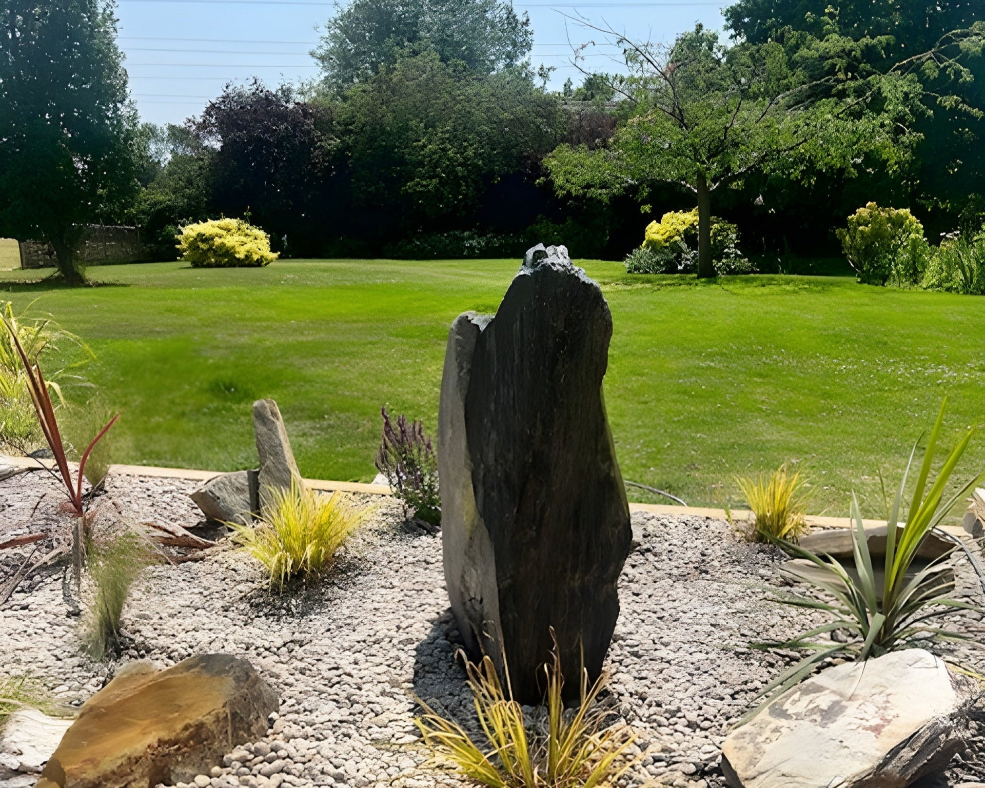 A landscaped garden featuring a tall, dark stone monument in the foreground with various plants and rock features. A lush, green lawn stretches into the background.
