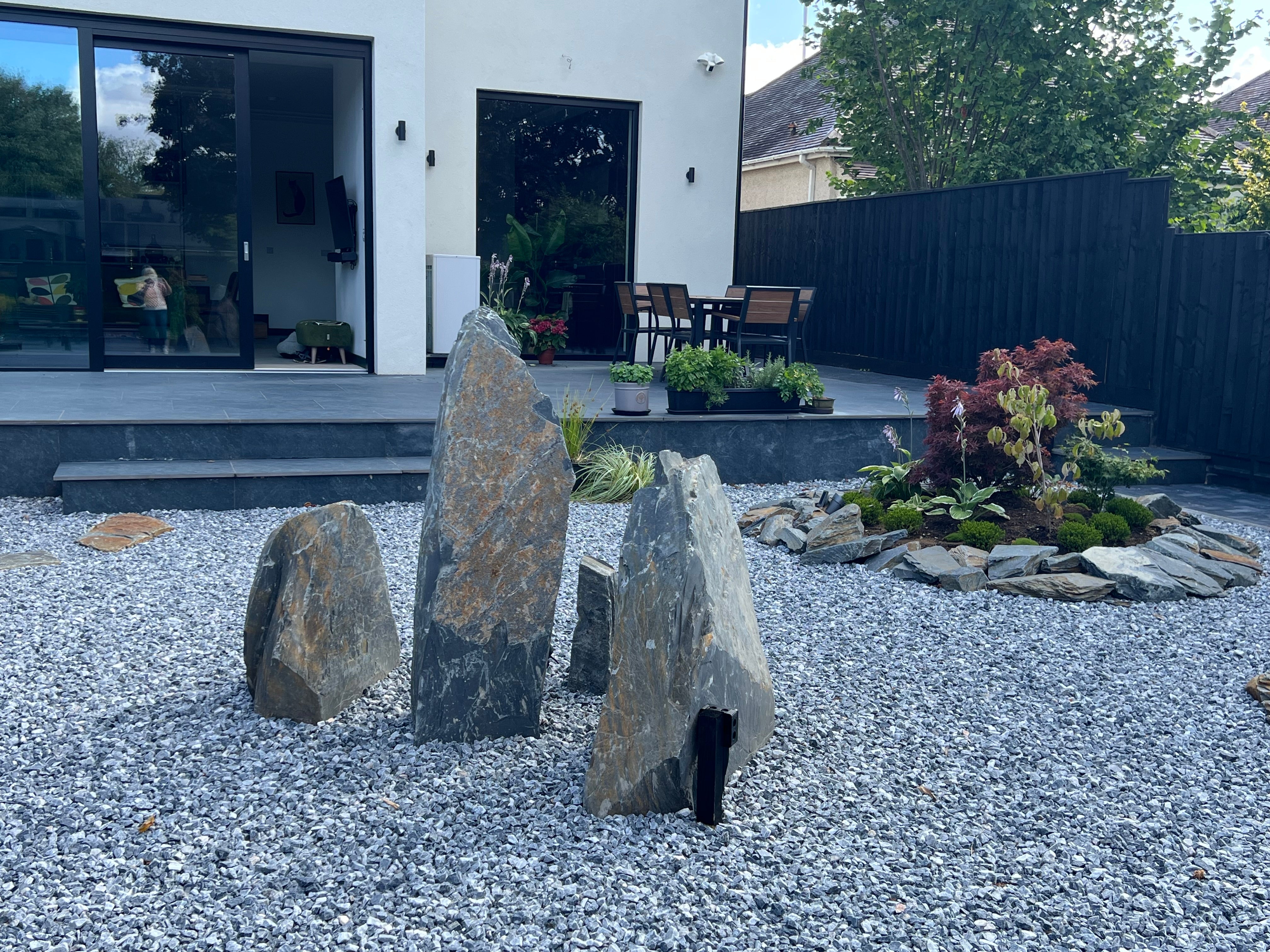A backyard zen garden features standing stones and a small planted island, covered with gray gravel, near a modern house.