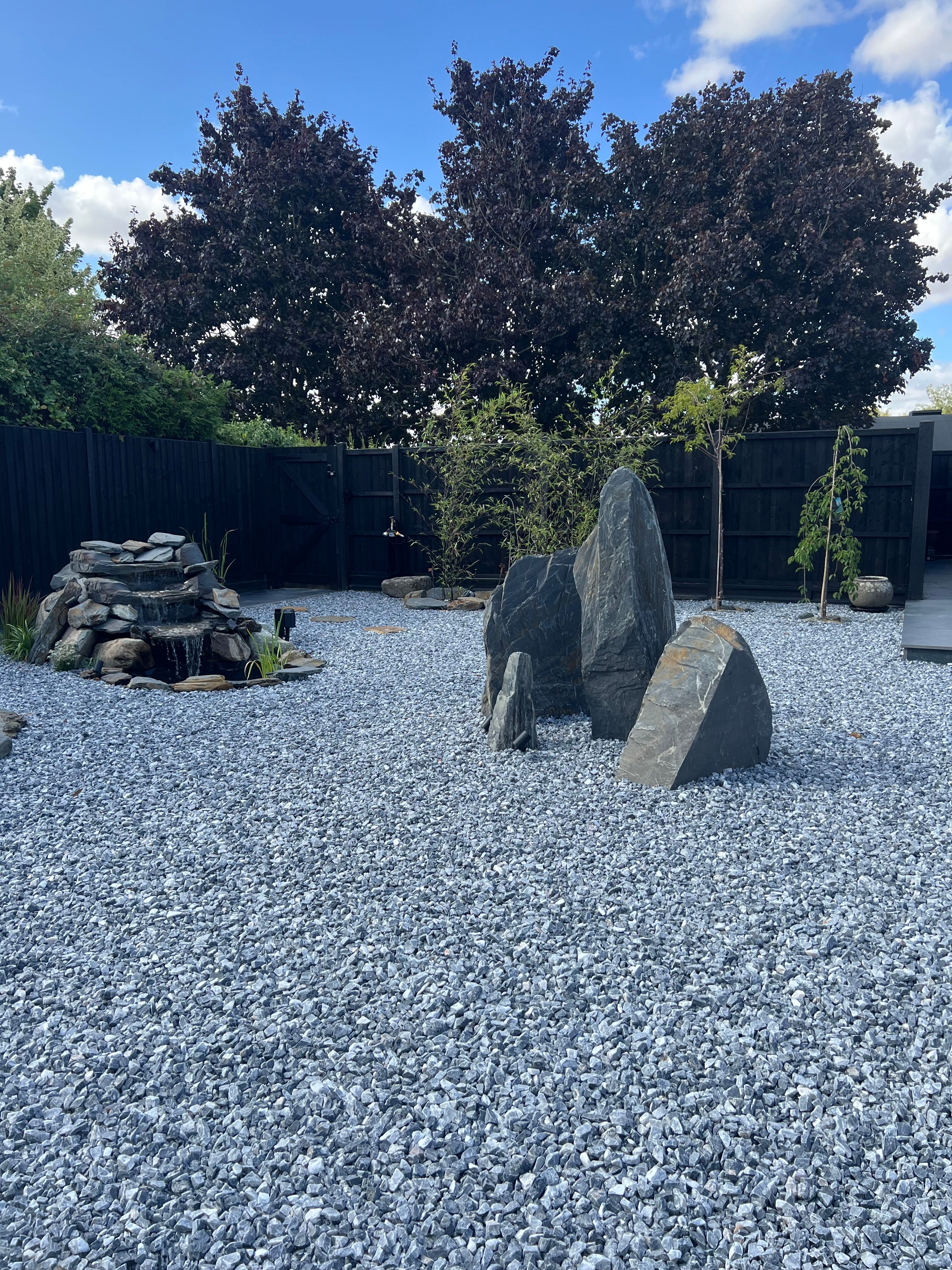 A serene garden with gray gravel, dark stones arranged as sculptures, a small waterfall, and dark wood fencing under a blue sky.