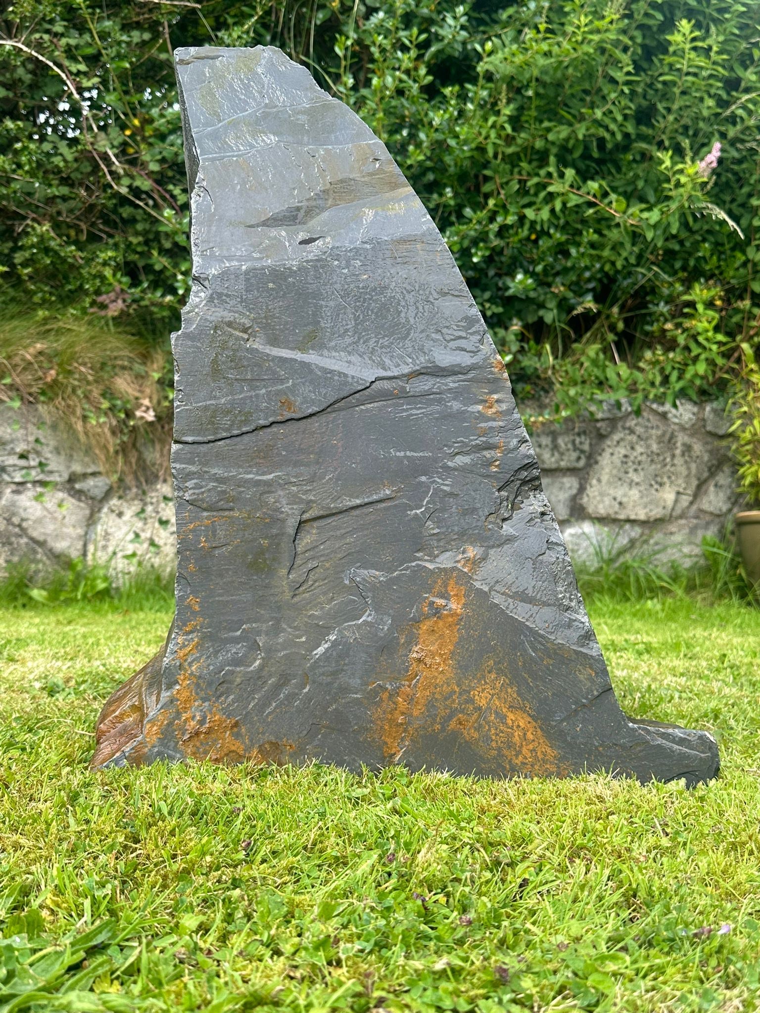 Grey slate monolith in the shape of a shark fin, with rust-colored weathering, set in green grass.
