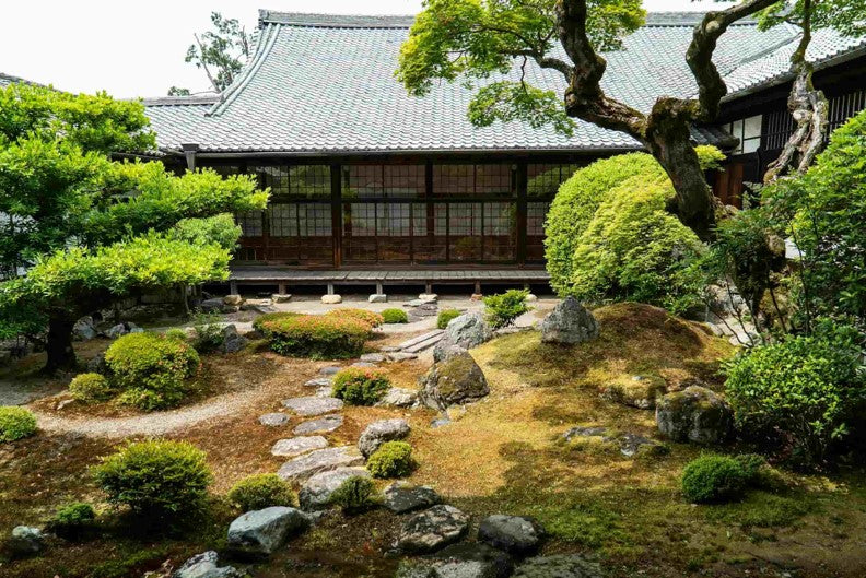 A Japanese garden with a wooden building in the background, featuring carefully placed stones and pruned trees. The scene is tranquil and peaceful.
