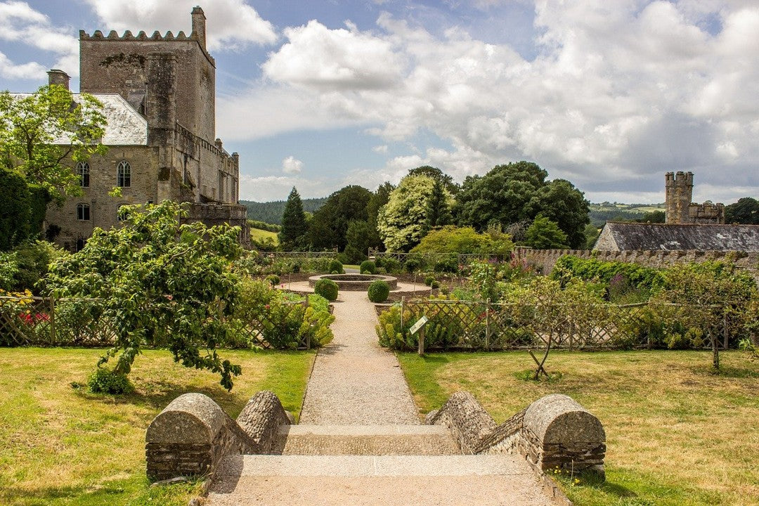 A castle with a surrounding formal garden is captured in a sunny, outdoor shot. Stone steps lead toward a garden path and lush greenery.
