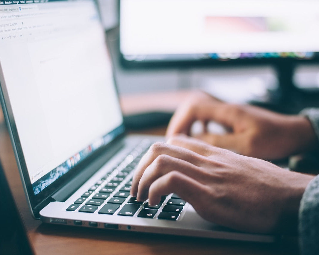 A person types on a laptop keyboard. A second monitor stands in the background. The laptop screen displays a Google Docs page.