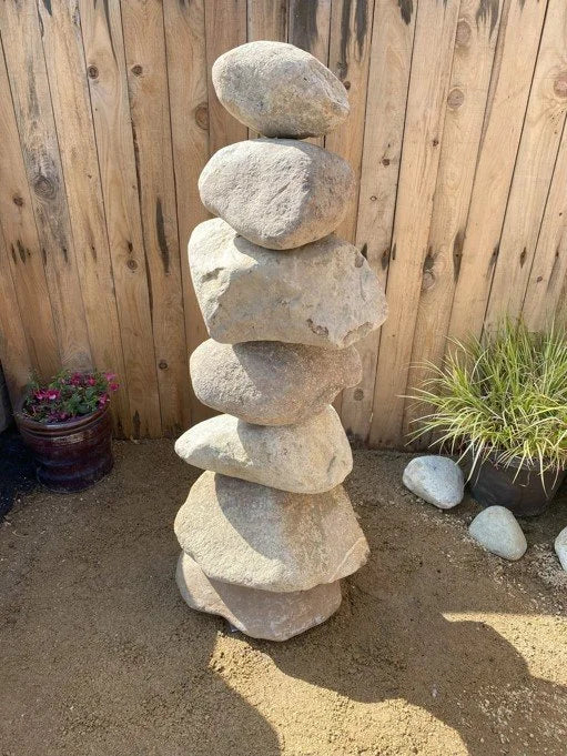 Large stacked stone pebble tower from unknown vendor, standing in sand next to potted plants.