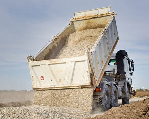 A white dump truck tilts backward, unloading gravel onto a dirt road under a bright sky.  A red and white sign is on the truck.