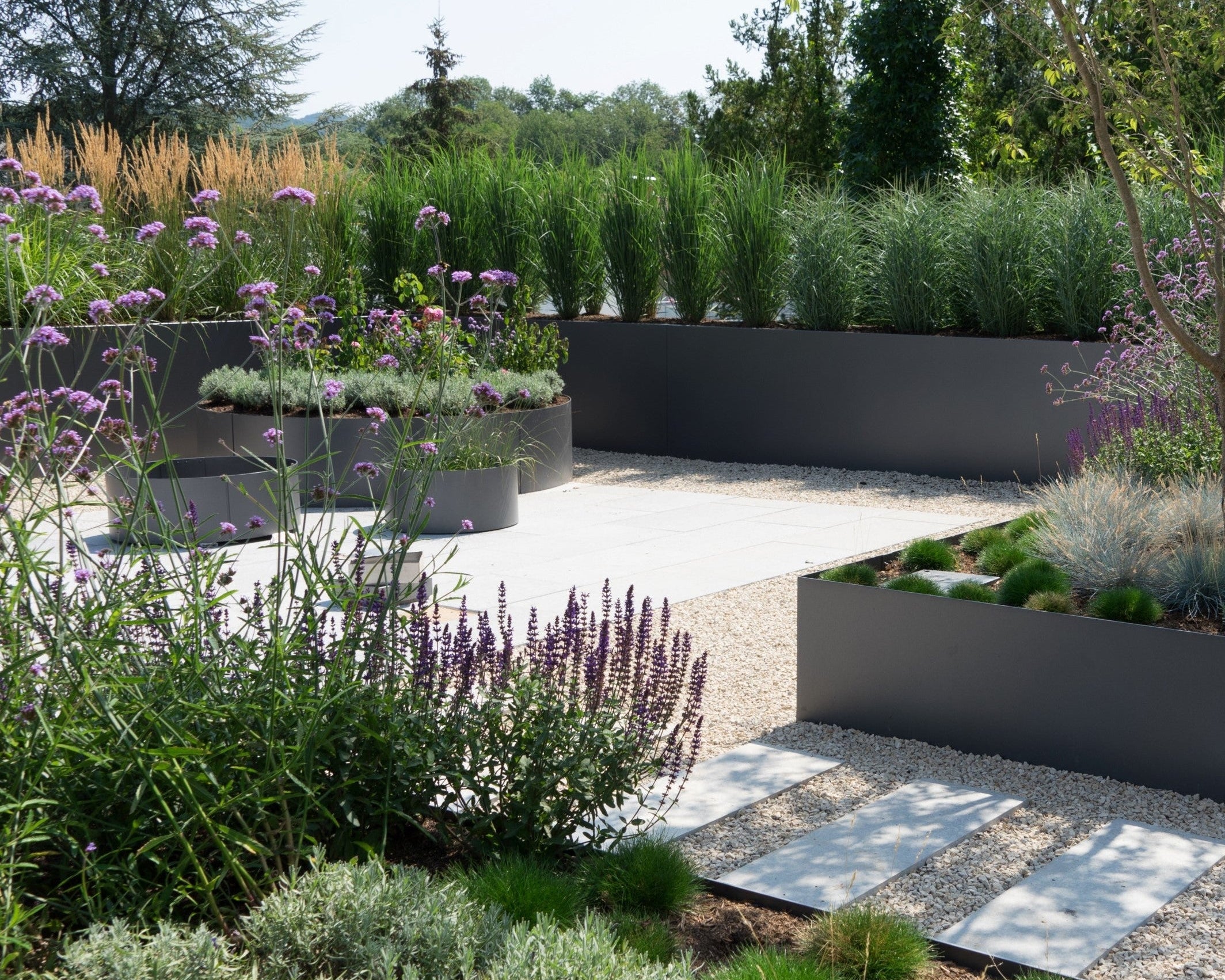 Modern garden with raised metal planters, grey stone pathways, and various green and purple plants under bright, natural light.
