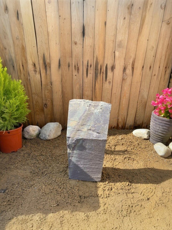 Slate monolith water feature with layered texture, standing in sand with plants beside a wood fence.