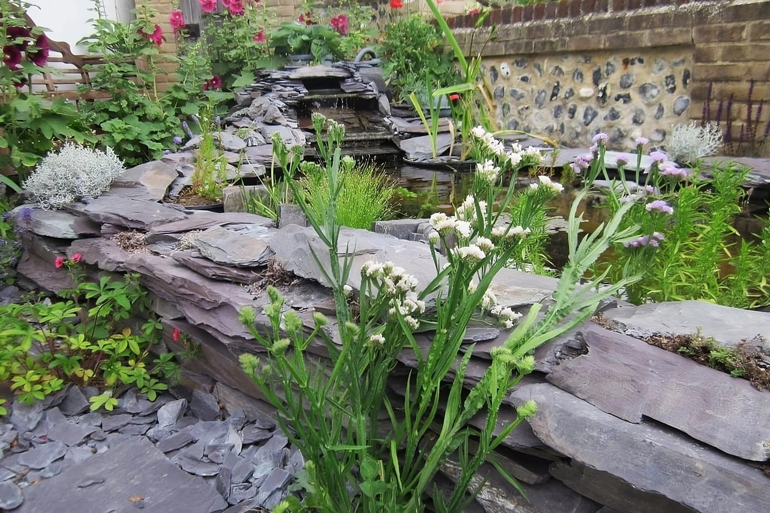 A stone garden with a small waterfall and pond, abundant greenery and flowers, showcasing a natural, peaceful atmosphere.