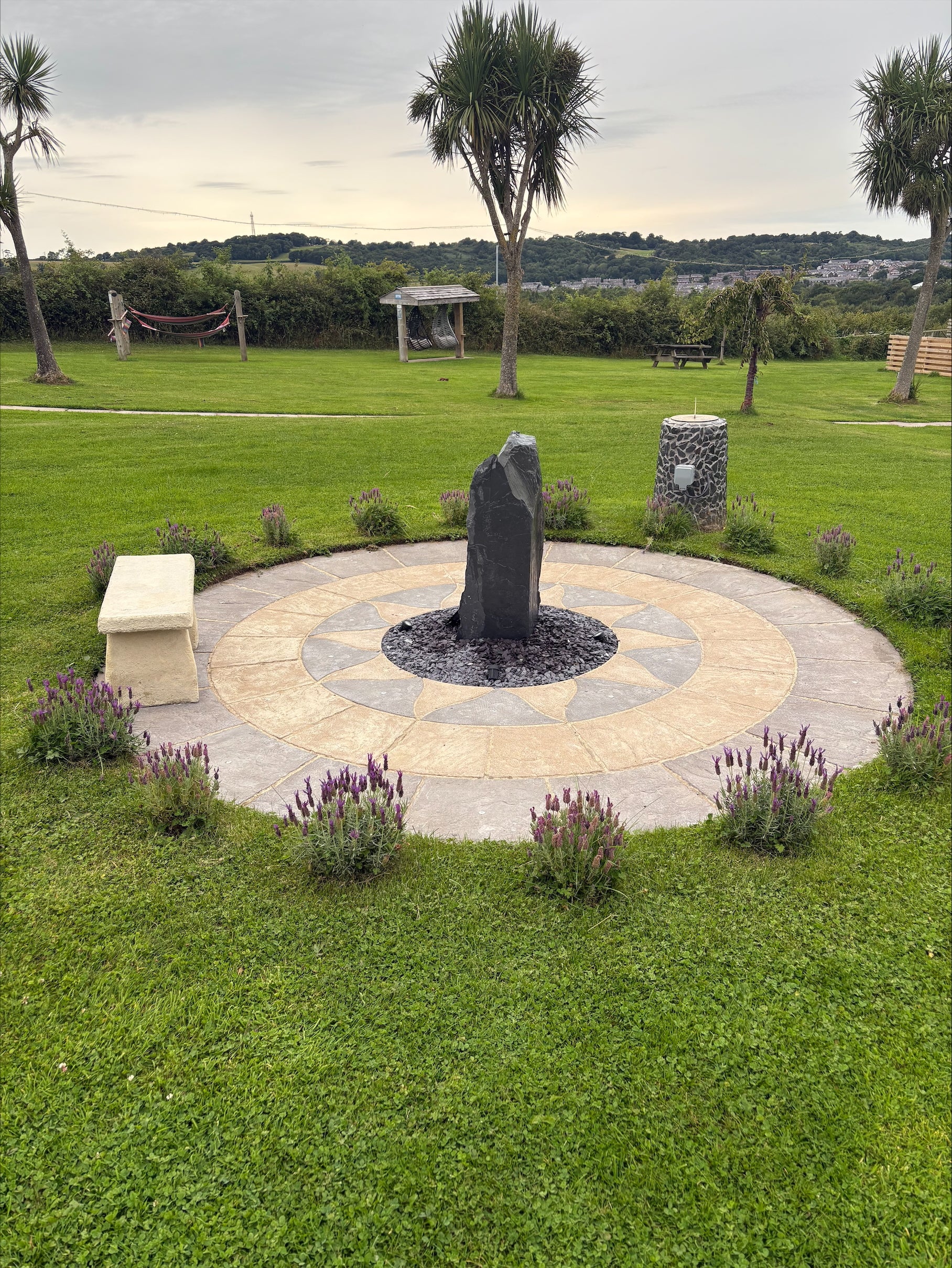 Outdoor scene with a circular stone water feature surrounded by lavender plants on a green lawn. There are trees, benches, and a hammock in the background.