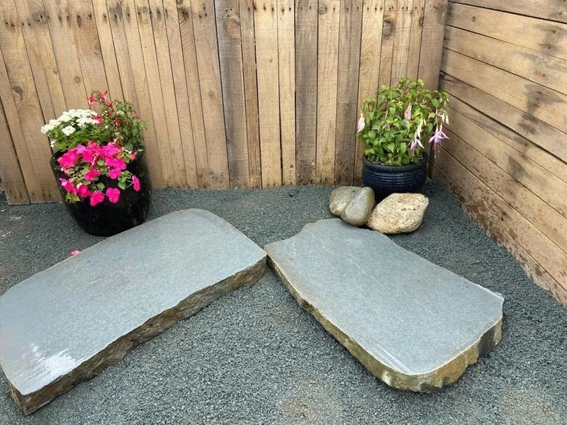 Sawn boulder modern stepping stones for ponds, set on gray gravel near potted plants and a wooden fence.
