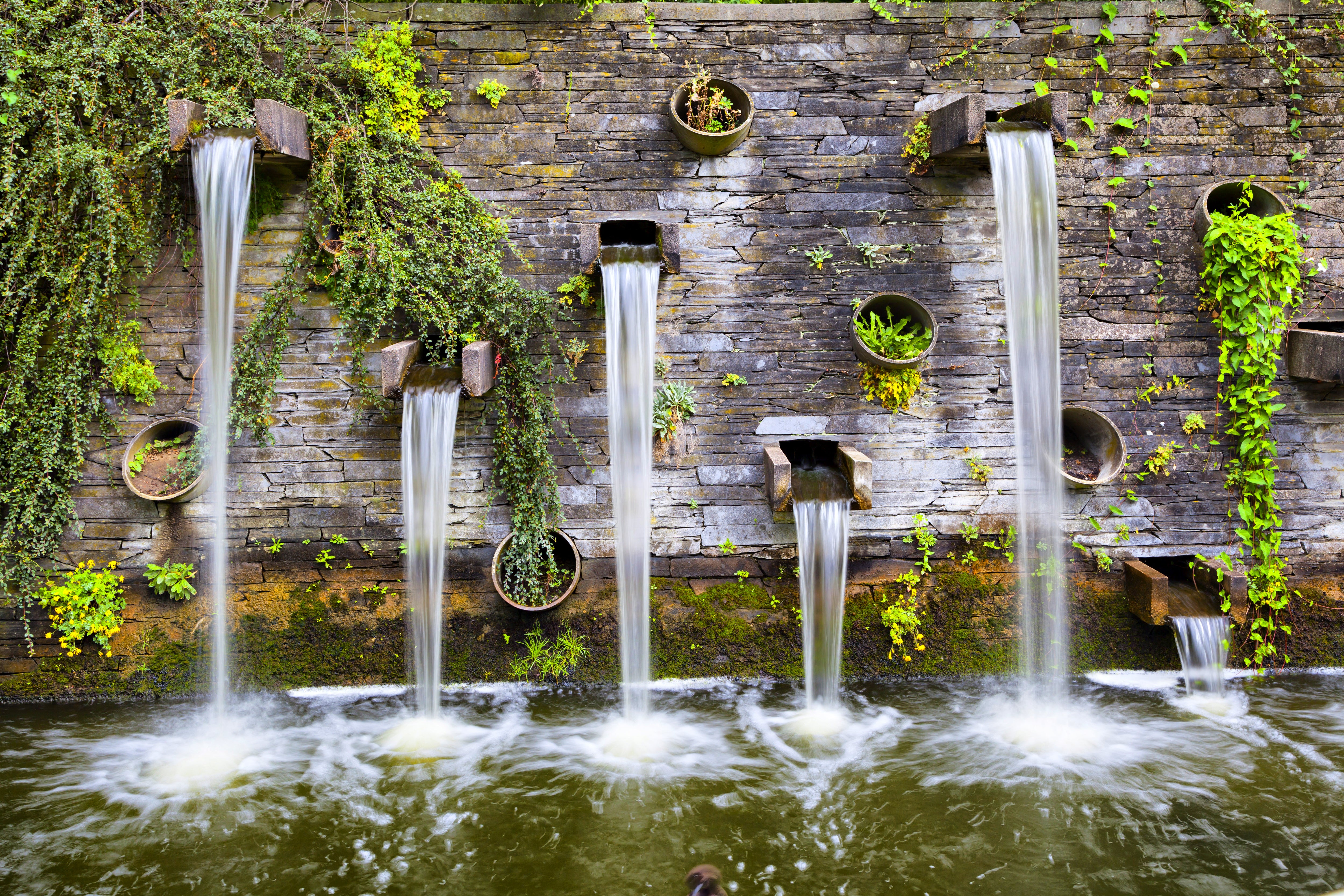 A serene water feature with multiple stone troughs pouring streams into a pond, adorned with lush greenery against a mossy stone wall.
