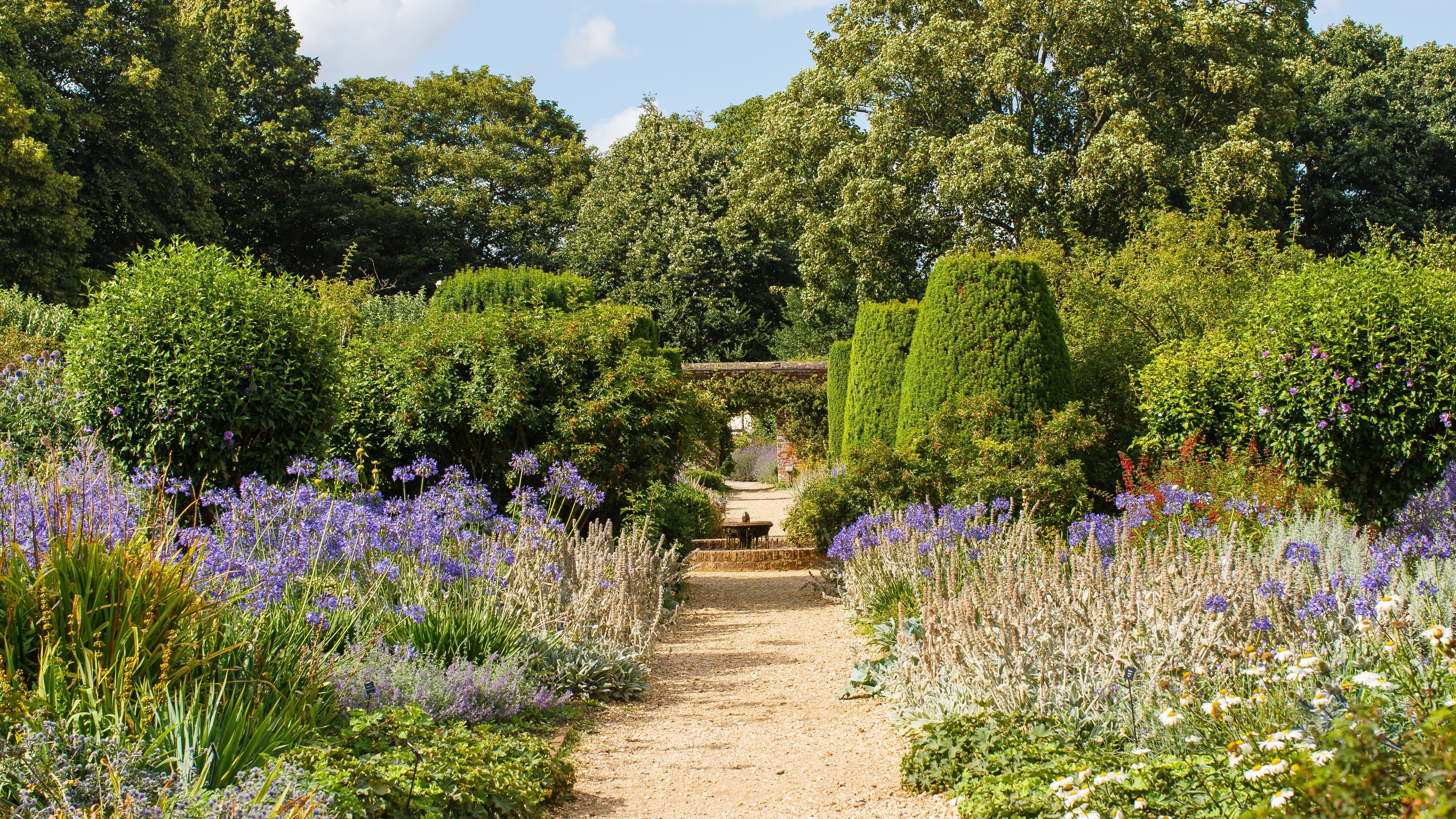 A garden path leads through lush greenery, with tall trees and a cloudy sky overhead, evoking a serene, natural ambiance.
