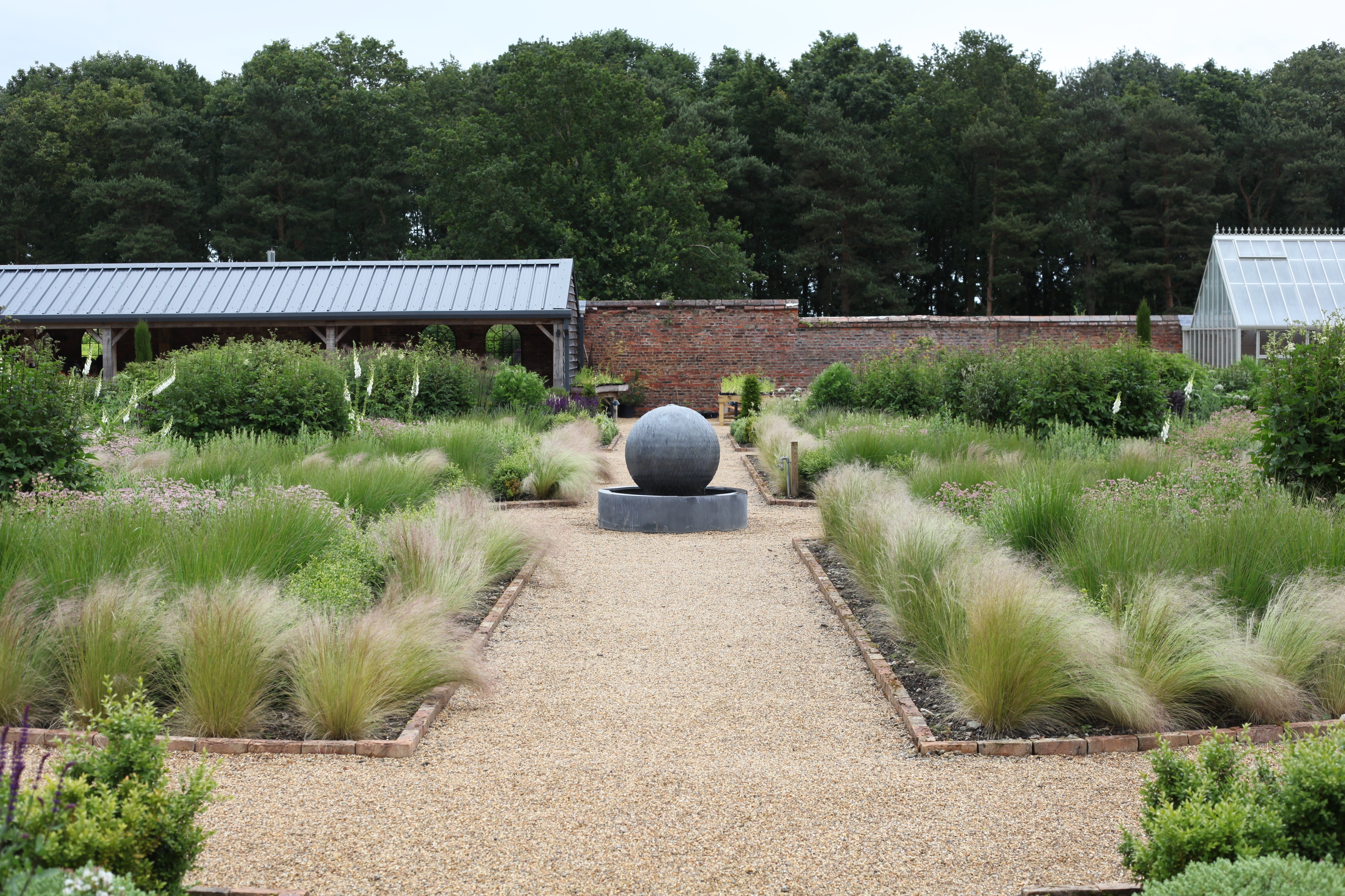 Formal garden with a gravel path leading to a stone sphere fountain, bordered by ornamental grasses and lush greenery. Brick walls and trees in the background.
