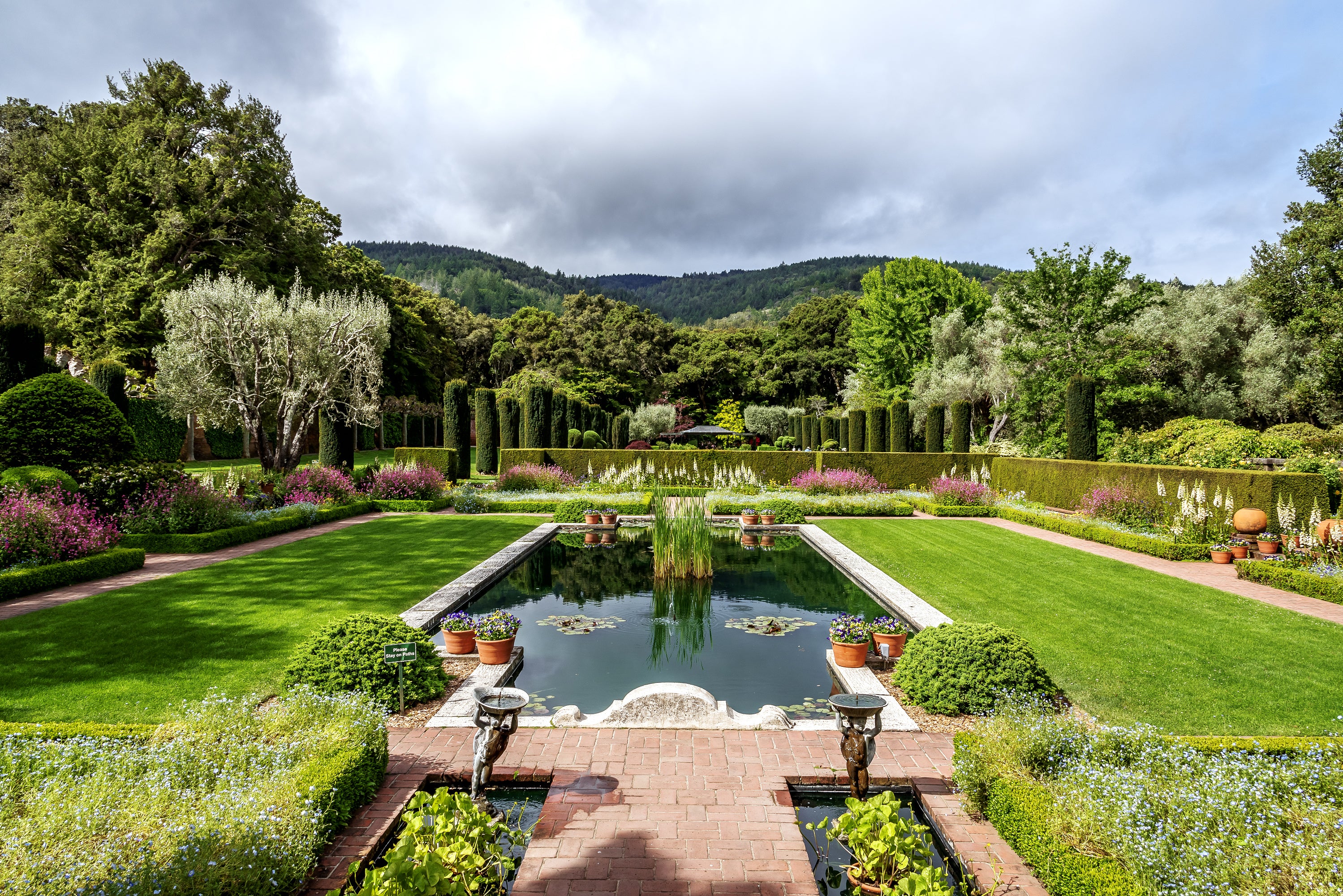 Formal garden with rectangular pond in foreground, green lawn, hedges, flowers, trees, and distant hills under cloudy skies.
