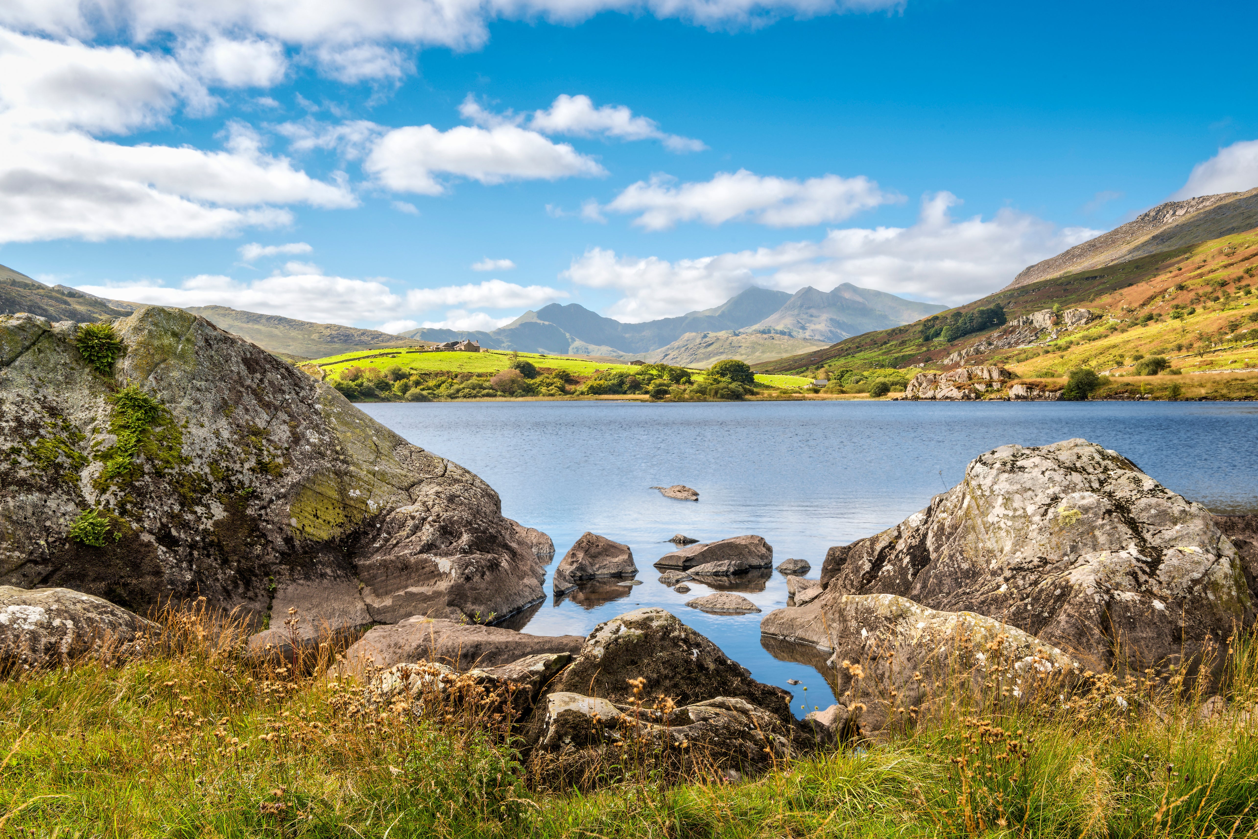 Scenic lake view framed by foreground rocks, reflecting clear blue sky with scattered clouds and distant mountains.
