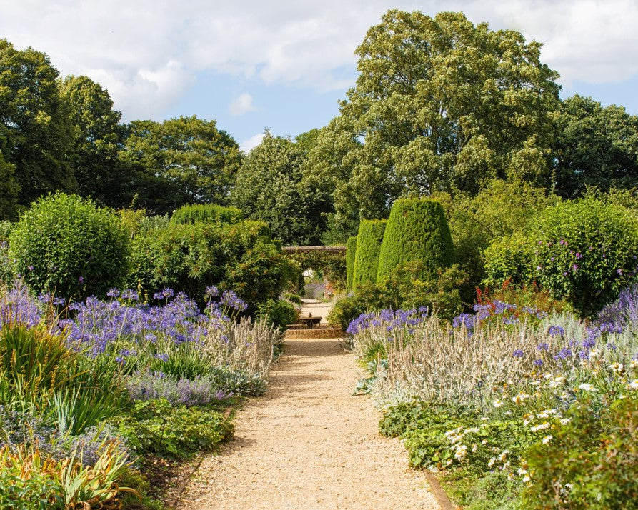 A lush garden path leads to a stone archway, lined with purple and white flowers and green shrubs. Tall trees stand in the background under a cloudy sky.
