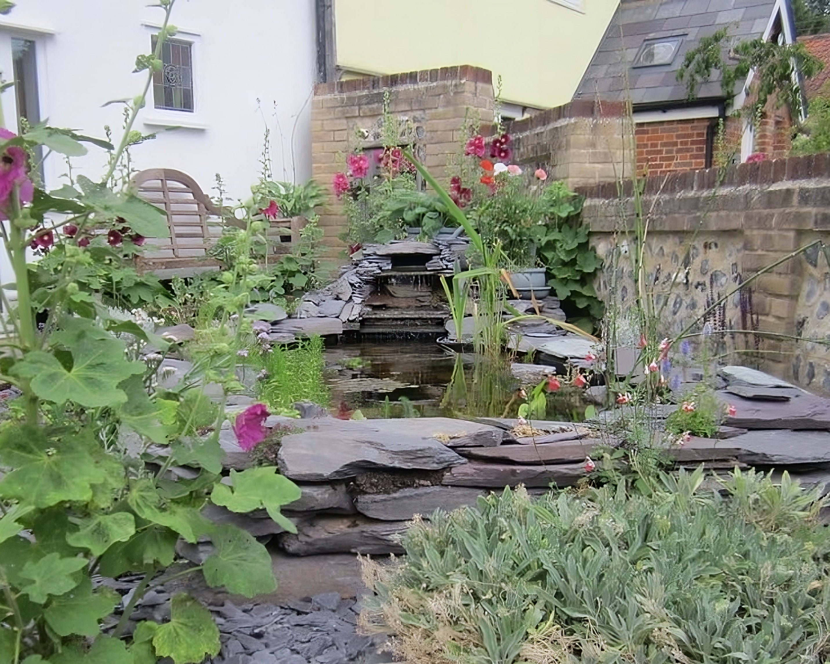 A lush garden with a slate-edged pond and small waterfall, surrounded by colorful flowers and greenery. Houses are visible in the background.