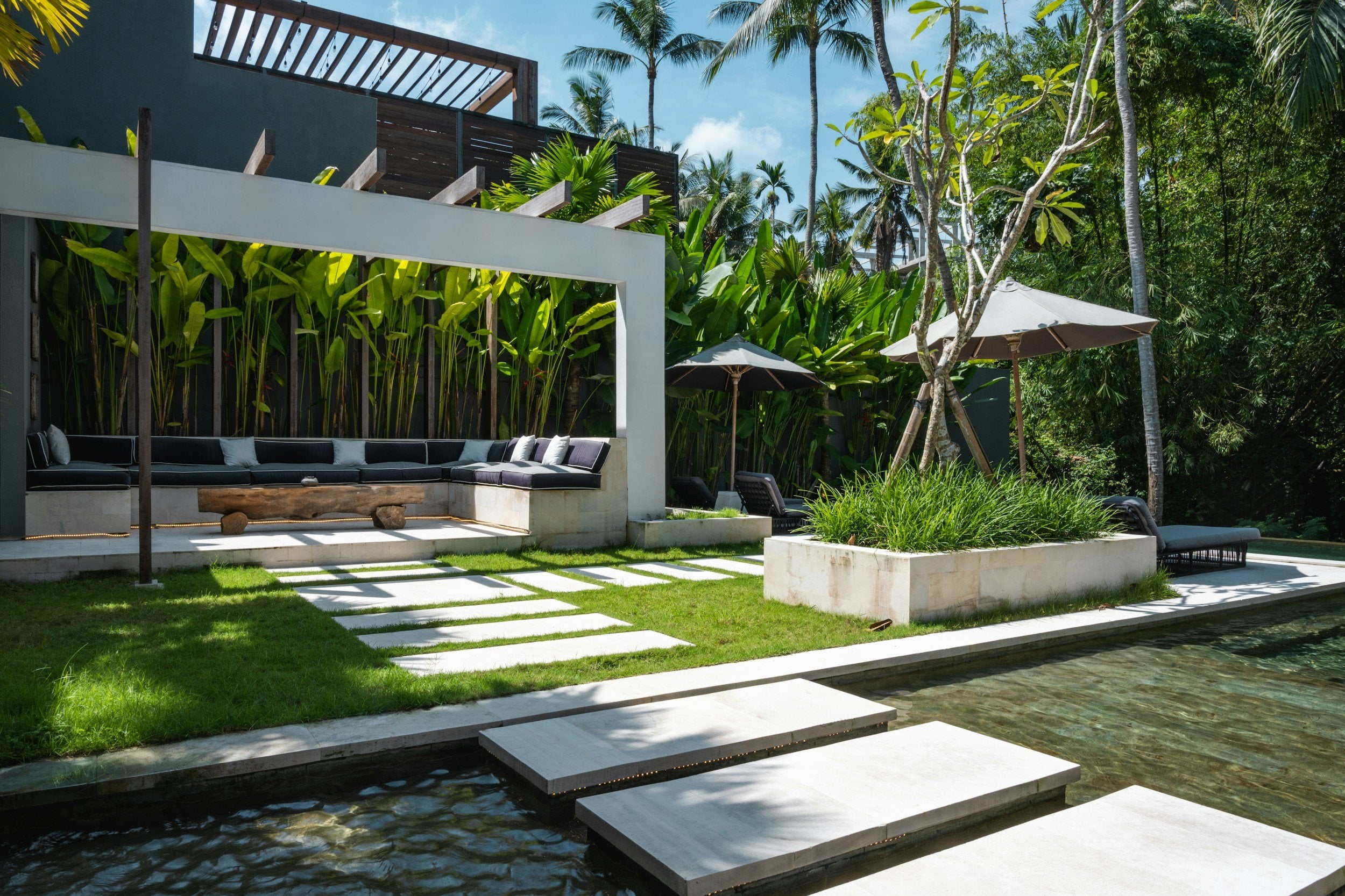 Outdoor lounge area with a white pergola, lush greenery, stone pathways, and stepping stones over clear water.