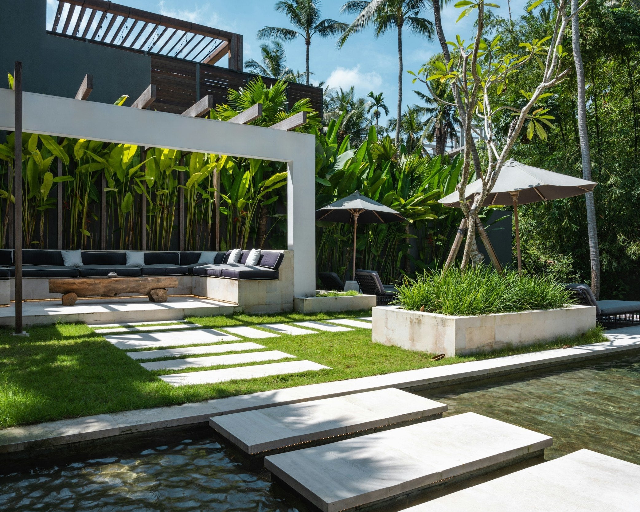 A modern outdoor lounge area with stone paths, green grass, black cushions, and tropical greenery, creating a tranquil and luxurious atmosphere.
